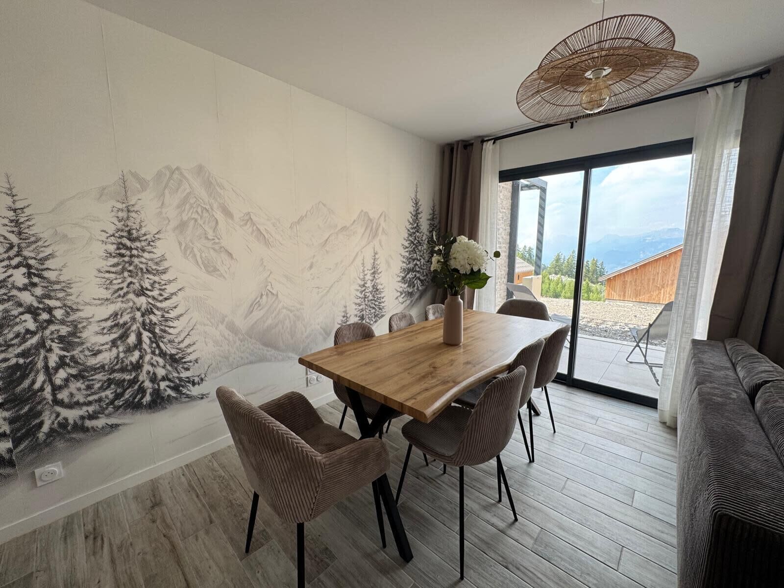 Un coin repas avec une table en bois, des chaises, un vase de fleurs et une fresque représentant un paysage de montagne sur le mur.