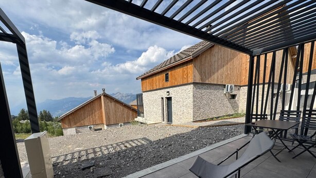 An outdoor seating area with tables and chairs, a wooden building, and a view of mountains. - Chalet Altéa - 7 à 8 pers - Neuf & Cosy - 300 m des Pistes (Risoul)
