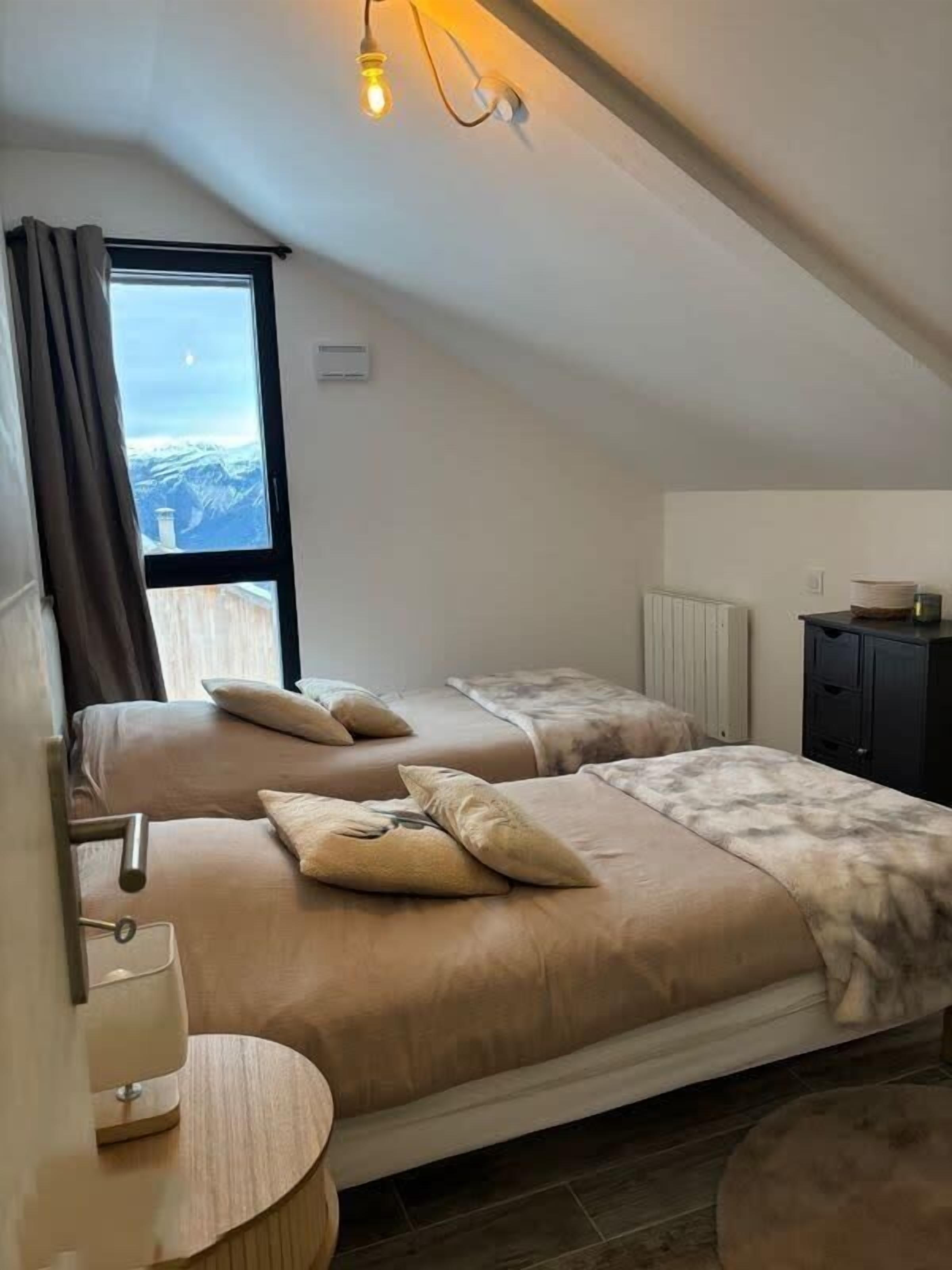 A bedroom with two beds, a window with curtains, a bedside table with a lamp, and a radiator.