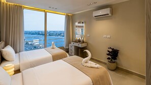 Quarto de hotel com duas camas, toucador com espelho, ar condicionado, vista para a cidade e um vaso de planta.