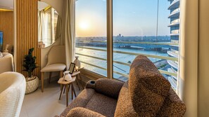 Ein modernes Hotelzimmer mit großem Fenster, einer Couch, einem Sessel, einem kleinen Tisch mit Blumenvase und einer Zimmerpflanze.