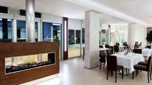 A modern dining area with tables set for guests, a central fireplace, and large windows offering views of the outdoors. - Vea Resort Hotel (Mercato San Severino)