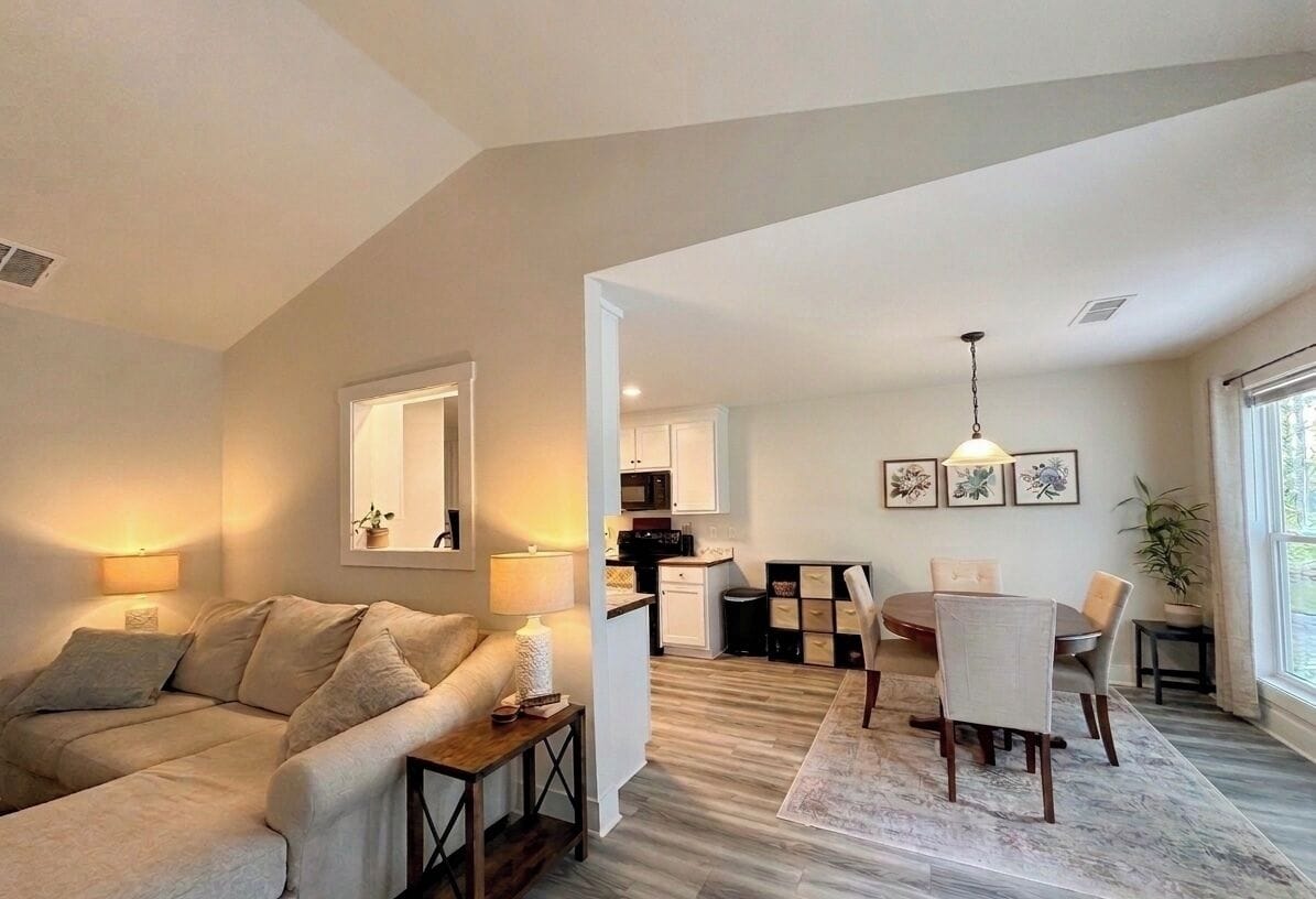 A living room with a sofa, a dining table with chairs, a kitchen area, and a large window.