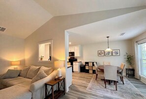 A living room with a sofa, a dining table with chairs, a kitchen area, and a large window. - Brand New Home! Modern Lake Retreat: 1 Mi to Water & Boat Parking (Hartwell)