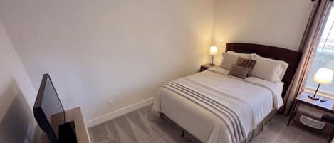 Una habitación de hotel con cama, mesita de noche con lámpara, televisión y una silla.