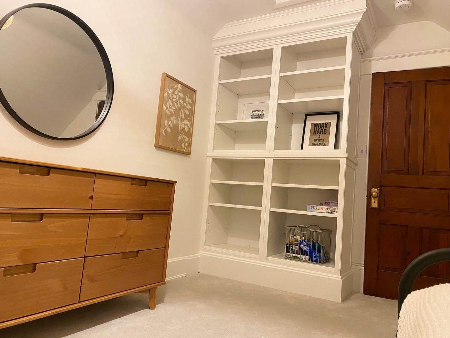 A room with a wooden dresser, a white shelving unit, a framed quote on the wall, and a door.