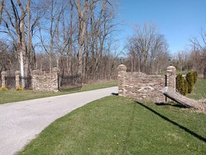 A paved pathway leading to a brick wall with a gate, surrounded by trees and grass. - Cozy 2-bedroom apartment in tranquil Birch Run with AC (Birch Run)