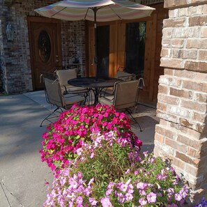 An outdoor patio area with a brick wall, a table with chairs, and a flower bed with pink flowers. - Cozy 2-bedroom apartment in tranquil Birch Run with AC (Birch Run)