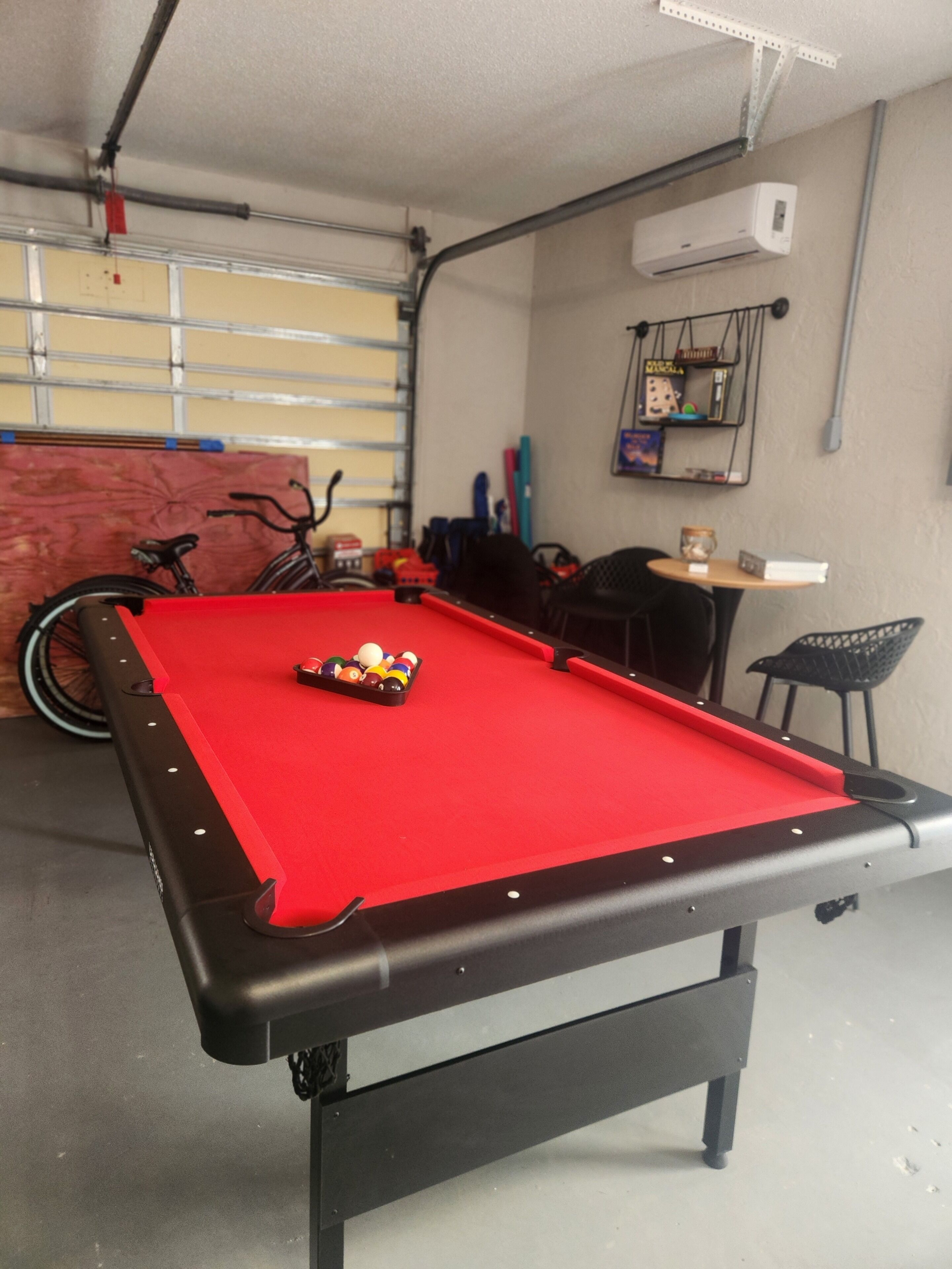 Une table de billard avec des billes, un vélo et une chaise dans une pièce dotée d’une porte de garage et d’un appareil de climatisation.