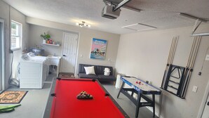 Uma mesa de bilhar em um ambiente com projetor no teto, um sofá, uma pequena mesa com suporte para tacos de bilhar e um pôster na parede.