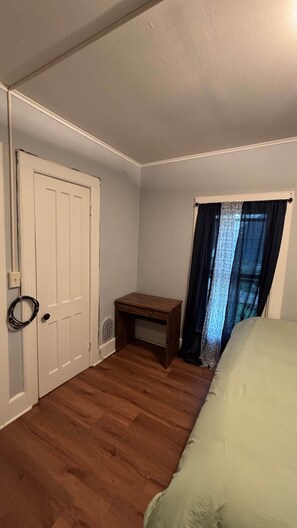 A bedroom with a wooden floor, a bed, a nightstand, a door, and a window with curtains. - Cozy Oneonta Getaway (Oneonta)