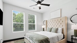 A bedroom with a bed, two windows, a ceiling fan, a bedside table with a lamp, and a wall-mounted mirror. - Modern Emerald Oasis 
Stylish Decatur Getaway (Decatur)