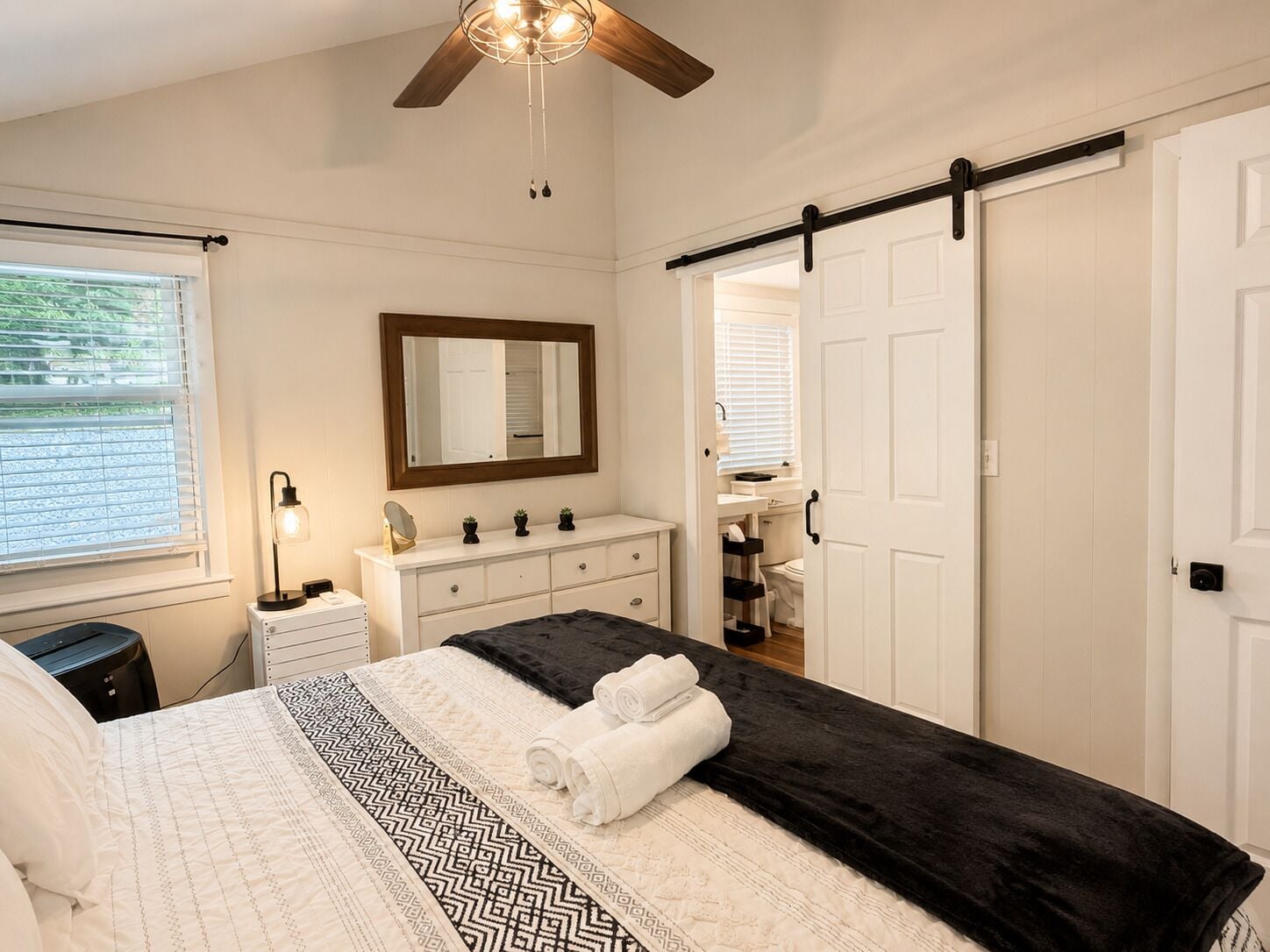 A bedroom with a bed, a dresser, a mirror, a ceiling fan, and a sliding door to a bathroom.