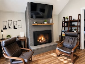 A living room with a fireplace, leather chairs, a TV mounted above the fireplace, and a bookshelf.