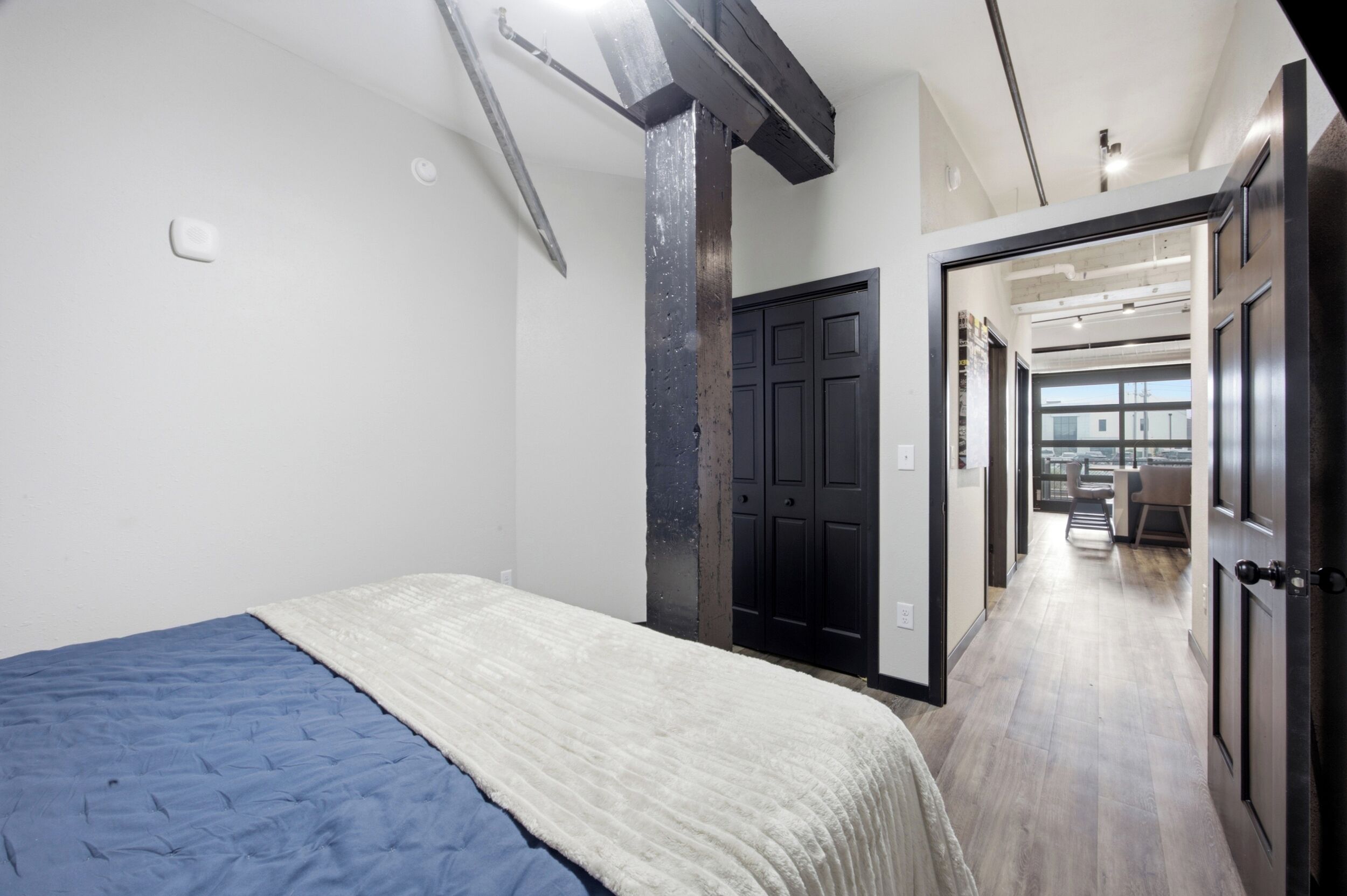 A bedroom with a bed, a door leading to a hallway, and a view of a cityscape through large windows.