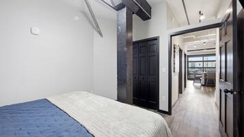 A bedroom with a bed, a door leading to a hallway, and a view of a cityscape through large windows.