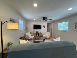 Un salon doté d’un ventilateur de plafond, d’un téléviseur à écran plat, de deux fauteuils blancs, d’une table basse et d’un canapé.