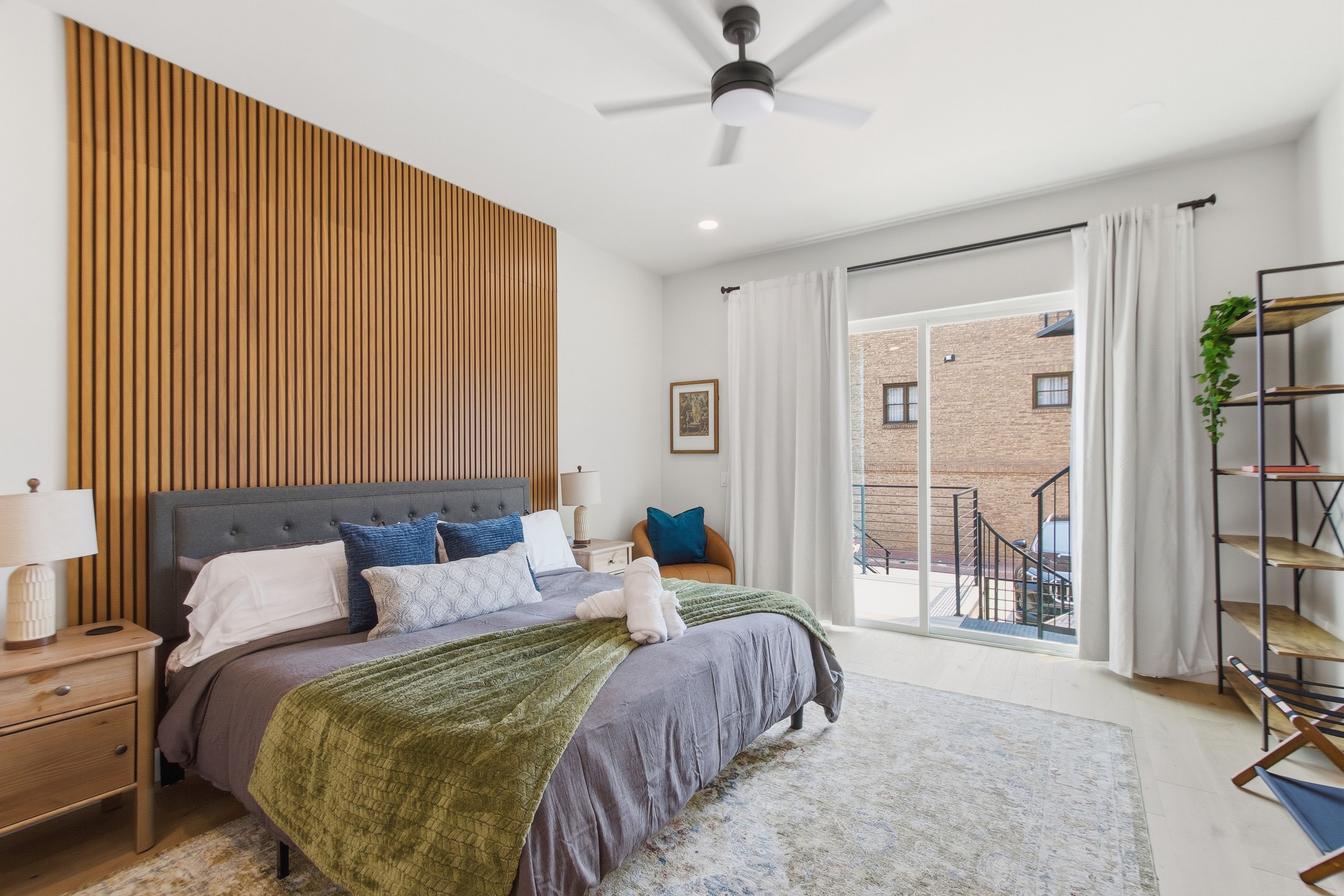 Ein modernes Schlafzimmer mit einem großen Bett, einem Deckenventilator und Blick auf ein Backsteinhaus durch das Fenster.