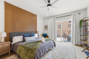 Ein modernes Schlafzimmer mit einem großen Bett, einem Deckenventilator und Blick auf ein Backsteinhaus durch das Fenster.