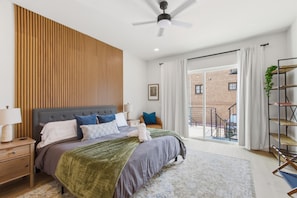 Ein modernes Schlafzimmer mit einem großen Bett, einem Deckenventilator und Blick auf ein Backsteinhaus durch das Fenster.
