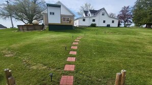 Un prato verde con un patio in legno che conduce a una casa con tetto verde e rivestimento bianco.