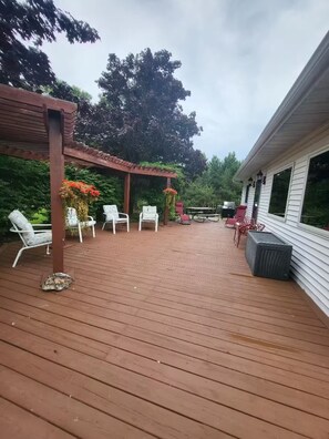 Una terraza de madera con mobiliario exterior, una pérgola y una casa al fondo.