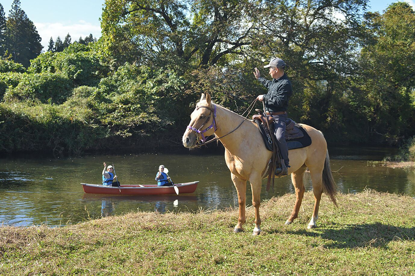 A horse with a rider standing on grass near a calm body of water, with a small boat and two people in it.
