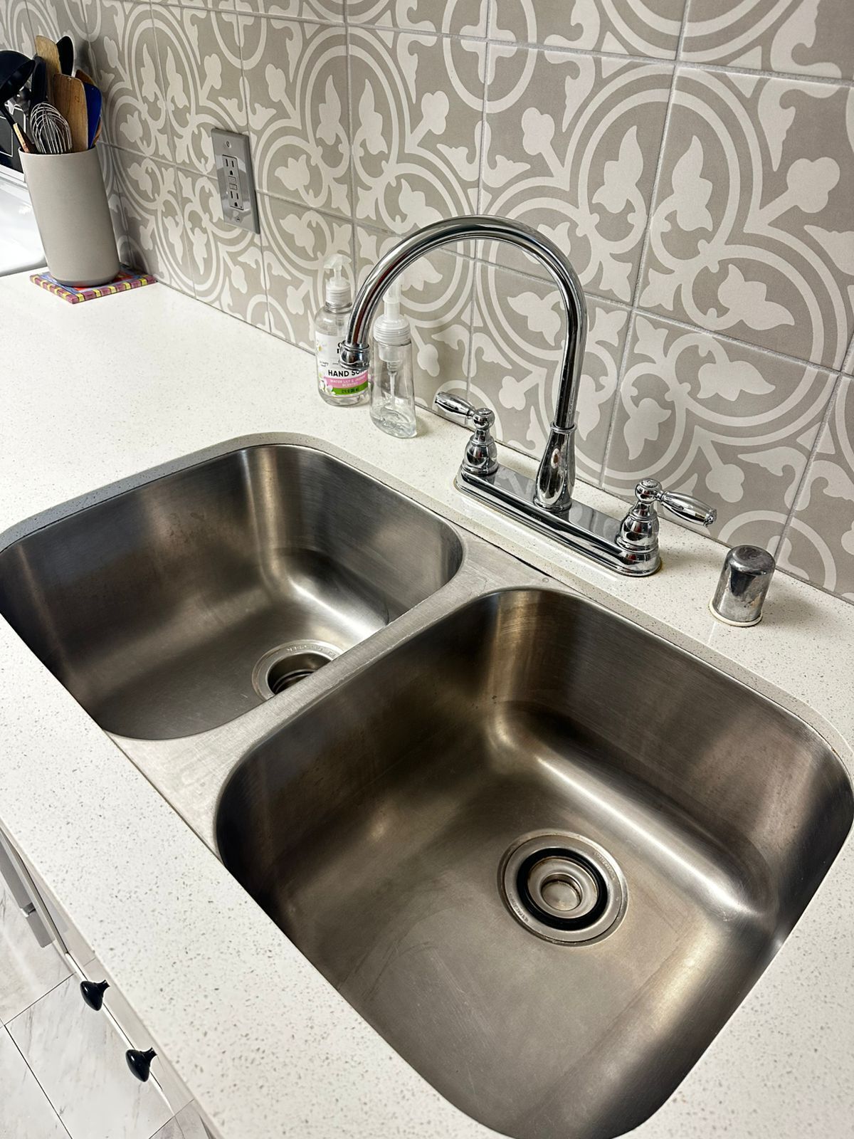 A stainless steel kitchen sink with a double faucet, a soap dispenser, and a utensil holder on the countertop.