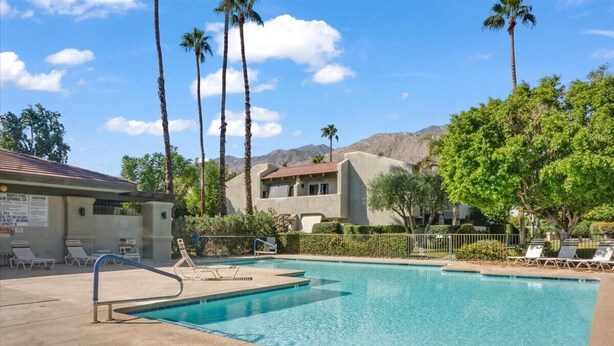 A swimming pool with lounge chairs, palm trees, and a building with a red roof. - Ground level condo with a large enclosed patio, steps from the pool (Palm Springs)