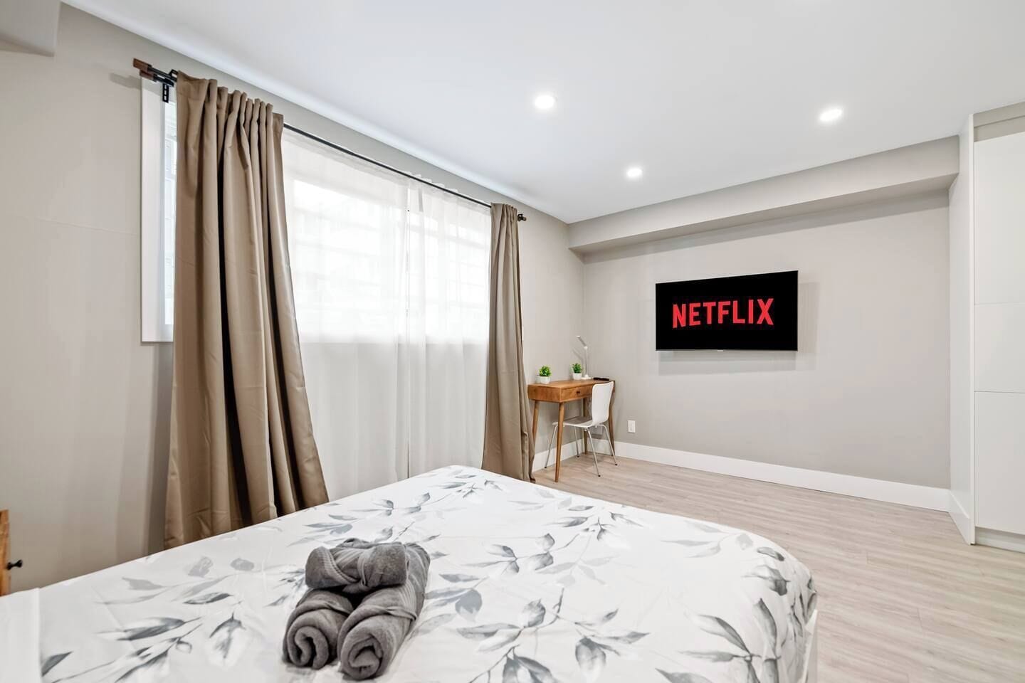 A hotel room with a bed, a desk, a chair, a TV screen displaying 'NETFLIX', and a window with curtains.