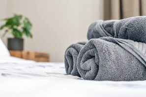 Ein ordentlich bezogenes Bett mit aufgerollten grauen Handtüchern und einer Blumentopf im Hintergrund.