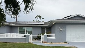 Une maison d’habitation avec un garage, une clôture blanche en bois et un palmier au premier plan.