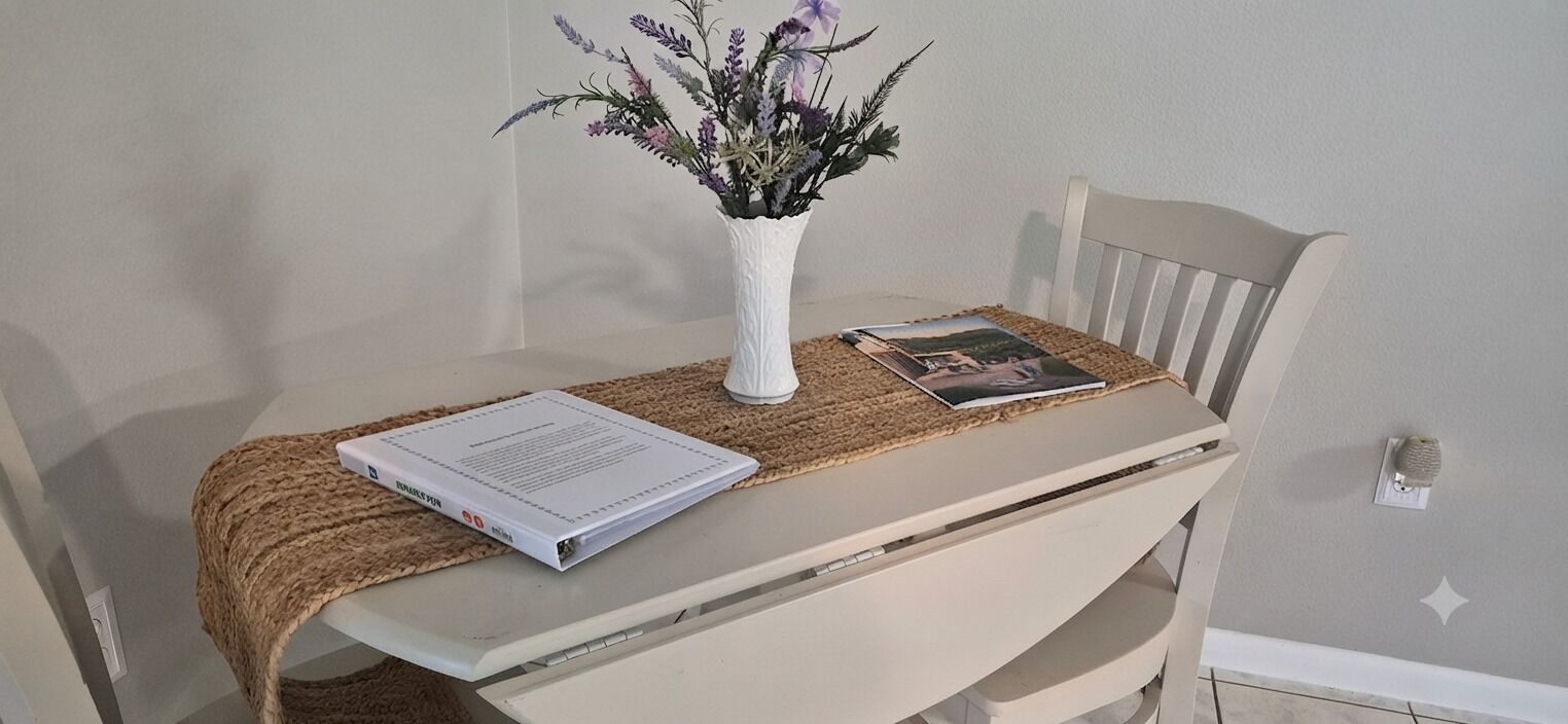 A white table with a woven placemat, a white vase with purple flowers, and a book on it.
