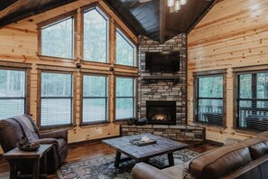 A log cabin interior with a stone fireplace, large windows, and leather furniture. - Secluded ~ Creek ~ Hot Tub ~ Patio ~ EV Charger (Broken Bow)