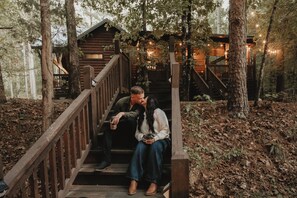 A wooden staircase leading to a cabin with a porch, surrounded by trees and a forest floor covered with leaves. - Secluded ~ Creek ~ Hot Tub ~ Patio ~ EV Charger (Broken Bow)