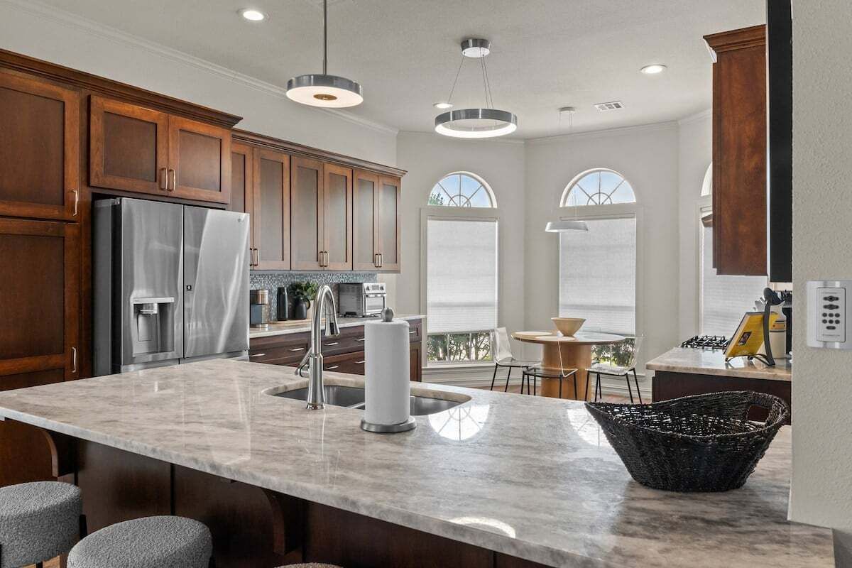 A modern kitchen with wooden cabinets, a stainless steel refrigerator, a marble island, and pendant lighting.