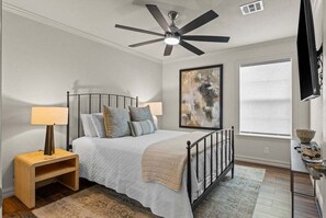 Ein Schlafzimmer mit Bett, Deckenventilator, Nachttisch, Lampe, einem Gemälde, einem Fenster und einem Fernseher.