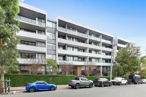 Un immeuble résidentiel à plusieurs étages, doté de balcons, avec des voitures garées et un ciel dégagé.