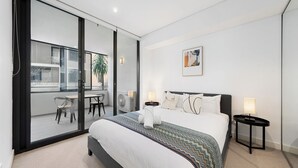 A modern bedroom with a large bed, a view of the outside balcony, and a small table with chairs. - Sleek 1BR Apartment | Home of Mortlake Ferry (Mortlake)