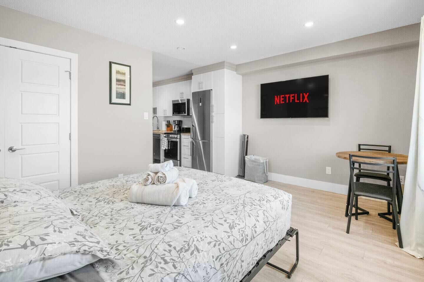A bedroom with a bed, a desk, a chair, a TV, and a kitchenette in the background.