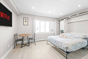 A bedroom with a bed, a small table with a chair, a window with curtains, and a wall-mounted TV. - Boutique-Style Studio Unit (Edmonton)