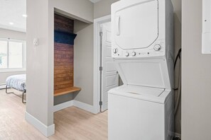 A laundry room with a washing machine and dryer, a wooden bench, and a bed in the adjacent room. - Boutique-Style Studio Unit (Edmonton)