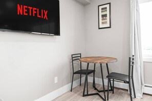 A room with a television displaying 'NETFLIX' in red letters, a round wooden table with two black chairs, and a framed picture on the wall. - Boutique-Style Studio Unit (Edmonton)