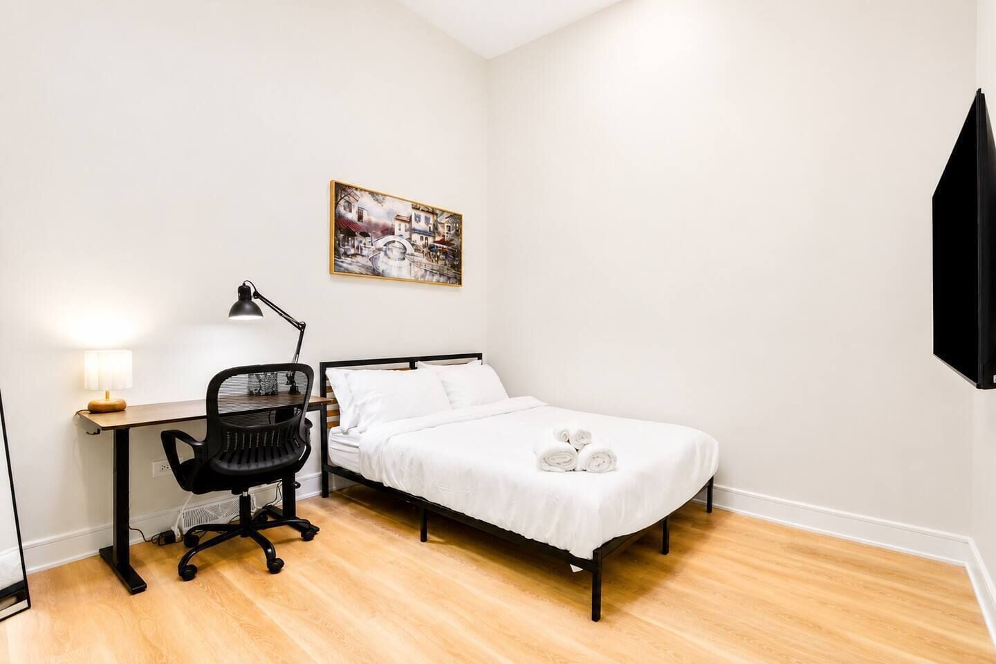 Un dormitorio con cama, escritorio con silla, lámpara y un cuadro en la pared.