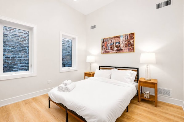 Quarto com cama, duas mesas de cabeceira, janela com tijolos aparentes e um quadro na parede.