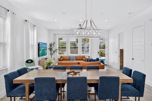 A spacious living room with a large wooden dining table surrounded by chairs, a leather sofa, and a television. - Destin Dreamin': 7 King Suites, Heated Pool, Golf Cart & Arcade Bunk Room (Miramar Beach)