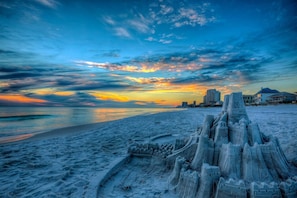 A beach with a sandcastle, a city skyline in the background, and a sunset. - Destin Dreamin': 7 King Suites, Heated Pool, Golf Cart & Arcade Bunk Room (Miramar Beach)