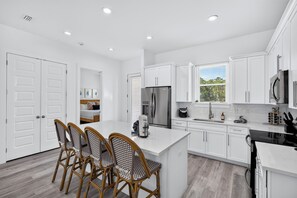 A modern kitchen with white cabinetry, a central island, and a dining area with wicker chairs. - Destin Dreamin': 7 King Suites, Heated Pool, Golf Cart & Arcade Bunk Room (Miramar Beach)