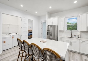 A modern kitchen with white cabinetry, a stainless steel refrigerator, and a dining area with wicker chairs. - Destin Dreamin': 7 King Suites, Heated Pool, Golf Cart & Arcade Bunk Room (Miramar Beach)