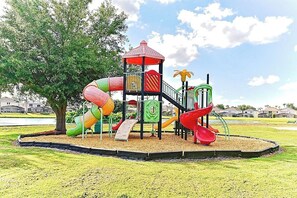 Ein Spielplatz mit Rutschen und einer Kletterkonstruktion, umgeben von Gras und Bäumen.